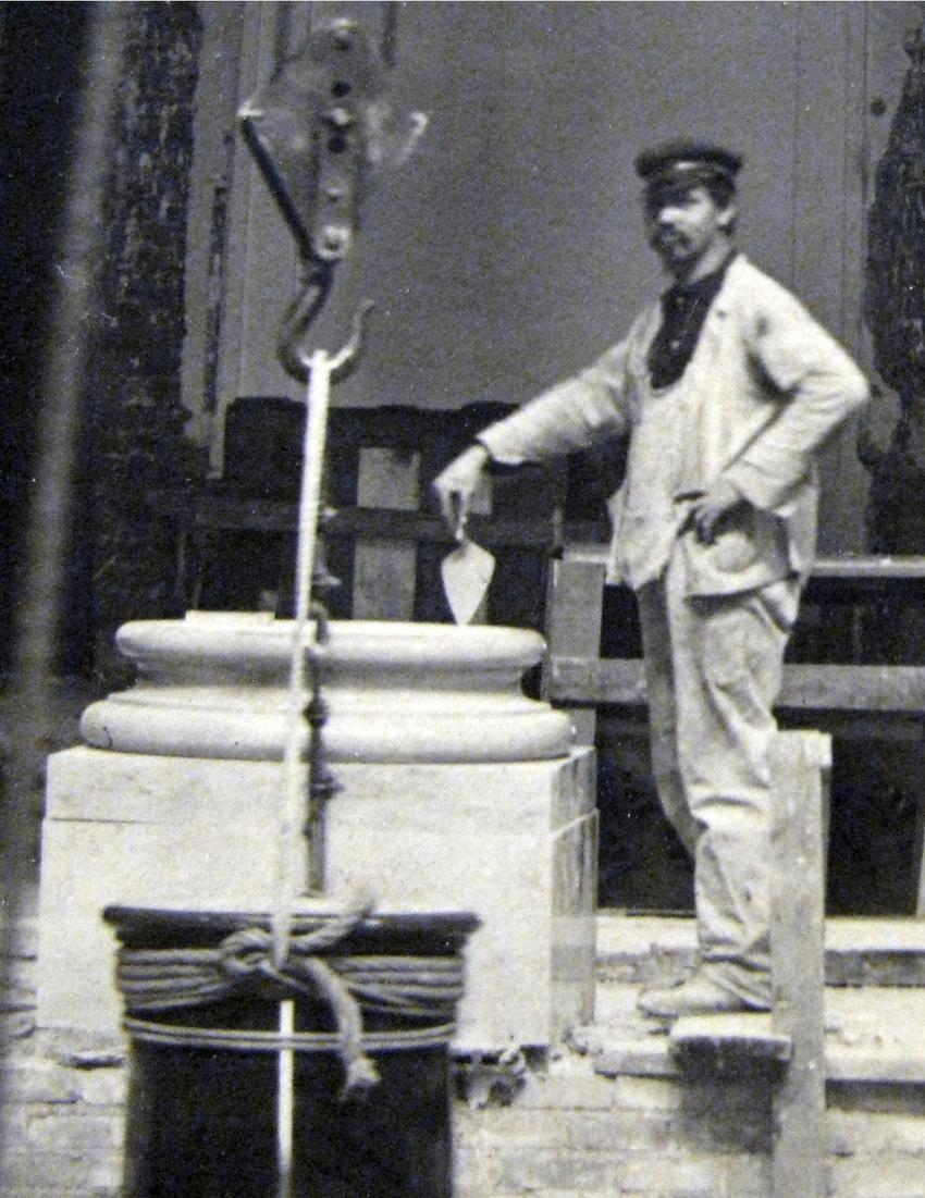 A stone mason stands next to a column base with his tools in hand.